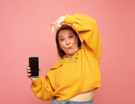 Téléchargez les photos : Femme adulte souriante portant un pull jaune montrant ou présentant un téléphone mobile sur fond rose - en image libre de droit