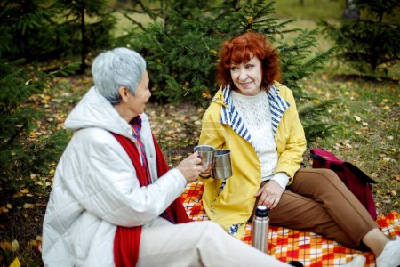 Téléchargez les photos : Deux vieilles amies sont assises sur une couverture à carreaux dans la forêt, buvant du café, parlant, s'amusant. Concept de style de vie et d'amitié féminine. - en image libre de droit