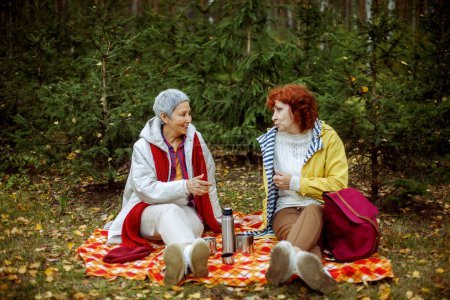 Téléchargez les photos : Deux femmes âgées sont assises sur une couverture à carreaux dans la forêt, buvant du café, parlant, s'amusant. Concept de style de vie et d'amitié féminine. - en image libre de droit