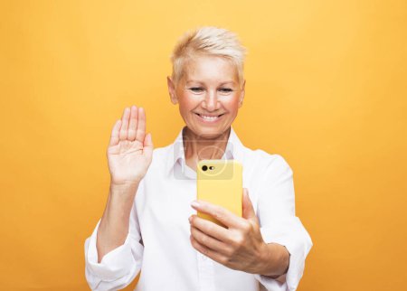 Téléchargez les photos : Mature attrayant femme avec les cheveux courts prendre un selfie sur fond jaune - en image libre de droit