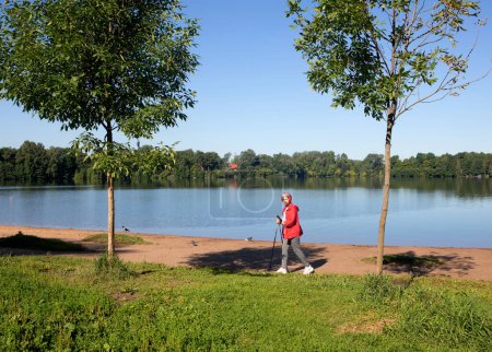 Téléchargez les photos : Femme aux cheveux gris portant une veste rouge marchant avec des bâtons de suivi sur la plage près du lac, jour d'été - en image libre de droit