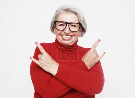 Foto de Portrait of funny senior gray-haired woman wearing red sweater and glasses doing a rock and roll symbol over white background - Imagen libre de derechos