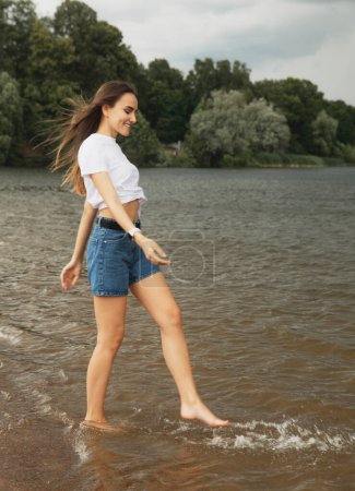 Foto de Joven alegre con el pelo largo camina por el lago descalzo. Salpicando en la orilla. Diversión y verano - Imagen libre de derechos
