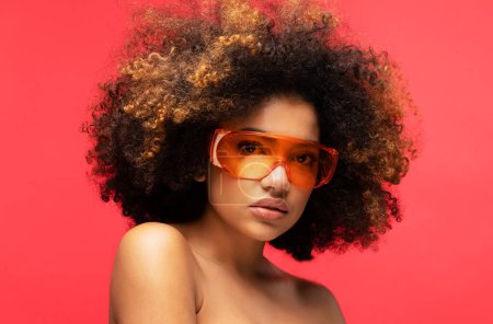 Foto de Mujer joven con gafas de sol aisladas en el estudio de fondo rojo. Emocionada mujer afroamericana. Primer plano retrato. - Imagen libre de derechos
