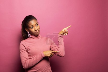 Foto de Mujer Afro asombrado señala lejos en el espacio de copia, discute promo increíble sobre fondo rosa - Imagen libre de derechos