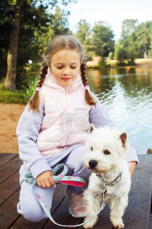Foto de A seven-year-old girl with a small white dog stands on a bridge near the lake and looks at the water. Friendship with pets. Happy childhood. - Imagen libre de derechos