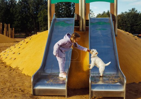 Photo for Seven-year-old girl playing in the playground with her white puppy, sunny summer day, lifestyle concept. - Royalty Free Image