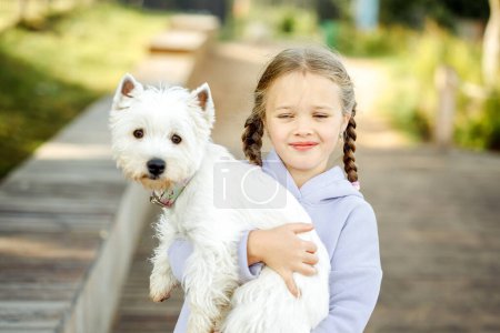 Téléchargez les photos : Une fille avec des nattes tient un petit chien blanc dans ses bras et sourit joyeusement. Jour d'été. - en image libre de droit