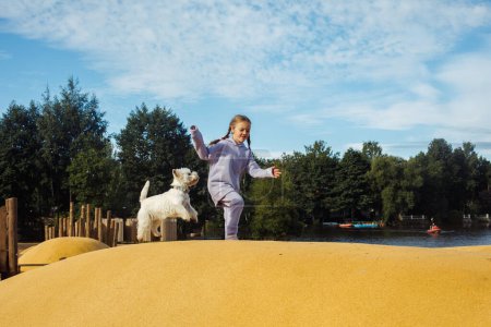 Foto de Niña de siete años jugando en el patio de recreo con su cachorro blanco, día de verano soleado, concepto de estilo de vida. - Imagen libre de derechos