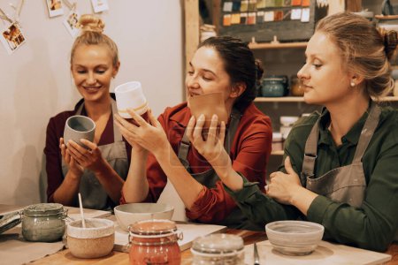 Téléchargez les photos : Une entreprise de trois jolies jeunes amies fabrique des tasses en céramique dans un atelier de poterie. Amusez-vous bien.. - en image libre de droit