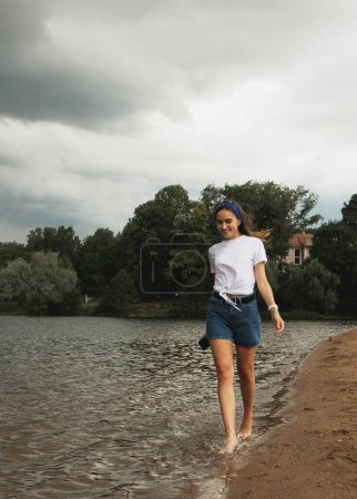 Foto de Joven alegre con el pelo largo camina por el lago descalzo. Salpicando en la orilla. Diversión y verano - Imagen libre de derechos