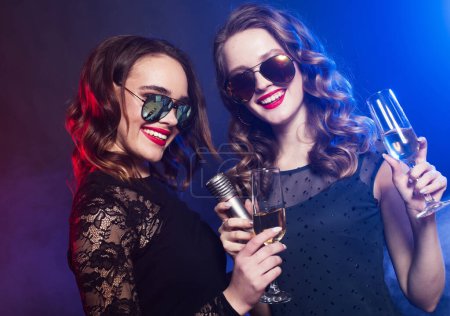 Téléchargez les photos : Style de vie, fête et concept de personnes : Deux jeunes jolies femmes portant une robe noire, tenant un verre de vin et chantant dans le microphone sur fond bleu - en image libre de droit