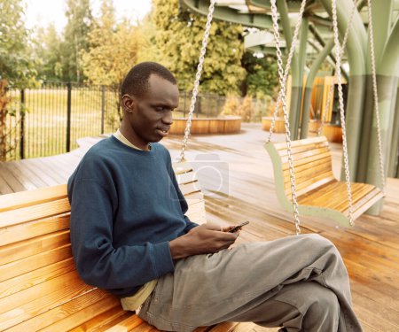 Téléchargez les photos : Jeune beau afro-américain assis sur un arbre dans le parc avec téléphone portable - en image libre de droit