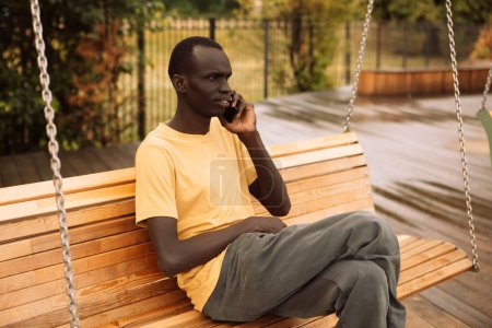 Foto de Joven guapo afroamericano sentado en un árbol en el parque con teléfono móvil - Imagen libre de derechos