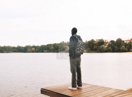 Téléchargez les photos : Jeune Afro-Américain se relaxant au parc près du lac. Jeune homme noir portant une chemise bleue à manches longues, avec un sac à dos, réfléchissant. Bonjour. Été. - en image libre de droit