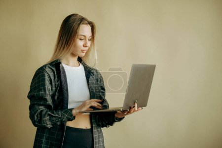 Téléchargez les photos : Souriant joyeux belle belle jeune femme 20s portant veste de base debout travaillant sur ordinateur portable PC regardant de côté isolé sur fond de mur couleur beige portrait studio - en image libre de droit