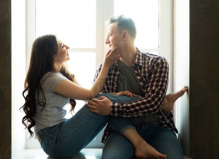 Photo for Lifestyle, family, love and people concept: Young couple hugging and kissing while sitting on the windowsill by the window - Royalty Free Image
