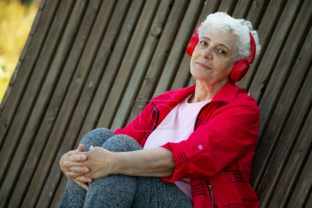 Foto de Una anciana con el pelo gris corto se sienta en un banco de madera en el parque y escucha música con auriculares, concepto de estilo de vida. - Imagen libre de derechos