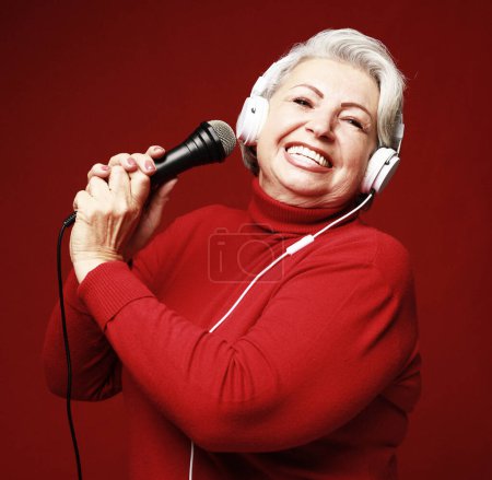 Téléchargez les photos : Vieille femme heureuse chantant avec microphone, s'amusant, exprimant son talent musical sur fond rouge - en image libre de droit