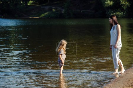 Téléchargez les photos : A five-year-old cheerful girl and her mother are walking by the lake, barefoot in the water, having fun on a summer day. Happy time. - en image libre de droit