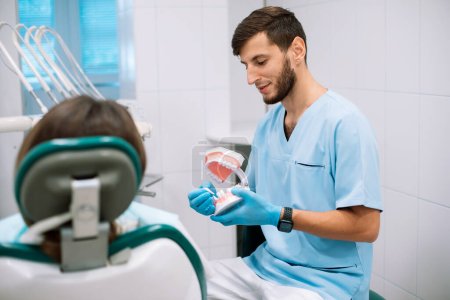 Männliche Zahnärztin zeigt dem Patienten Prothesen mit einer Halterung in der Hand. Falsche Zähne in den Händen des Folgearztes. Zahnprothetik. Falsche Zähne.