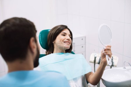 Foto de Mujer sonriente mirando su hermosa sonrisa sentada en el consultorio dental - Imagen libre de derechos