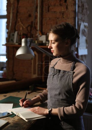 Téléchargez les photos : Jeune femme brune écrit au carnet en atelier de couture, concept fait main - en image libre de droit