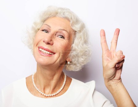 Photo for Elderly woman making selfie with peace sing and smilling. Portrait of expressive grandmother wearing casual over white background. - Royalty Free Image