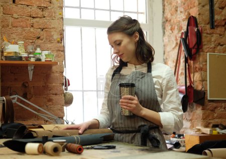 Téléchargez les photos : Femme travaille dans un studio de fabrication de sacs, découpe les détails et boit du café - en image libre de droit