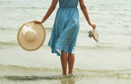 Foto de Imagen de una joven con un vestido azul caminando descalza en una playa y cuelga sus pies en el agua. Mujer joven en vestido blanco de verano con sombrero de paja mirando al cielo y al mar. Vista trasera. - Imagen libre de derechos