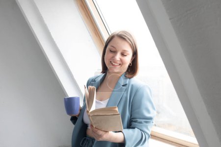 Photo for Smiling adult woman reading book and drinking coffee near window at home - Royalty Free Image