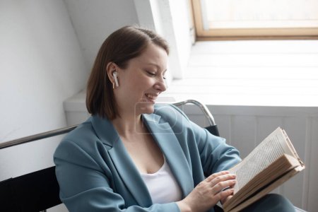 Foto de Mujer de mediana edad de moda relajado conpelo castaño claro sentado en silla moderna con libro cerca de la ventana, estilo de vida conept. - Imagen libre de derechos