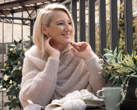 Téléchargez les photos : Une dame blonde âgée souriante s'assoit un matin d'été sur le balcon avec du tricot et une tasse de café - en image libre de droit