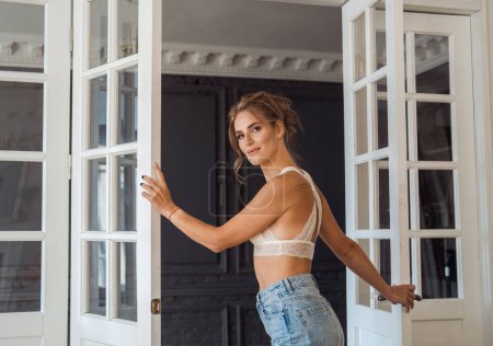 Photo for Charming blonde young woman in jeans and a lace top is standing in the living room - Royalty Free Image
