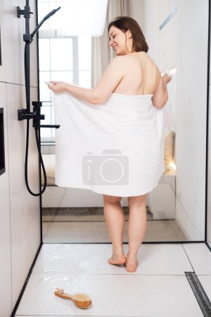 Photo for Adult plump woman grunts in a towel in the shower. Back view. - Royalty Free Image