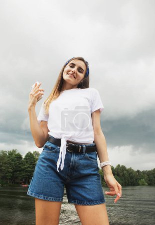 Foto de Mujer hermosa alegre en pantalones cortos y camiseta blanca es relajante junto al lago, disfrutando del día de verano - Imagen libre de derechos