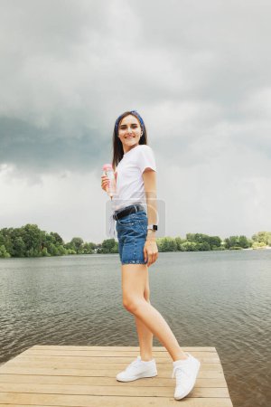 Photo for A cheerful young woman in shorts and a white t-shirt stands on a wooden bridge by the lake and enjoys a summer day. Lifestyle concept. - Royalty Free Image