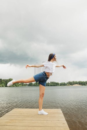 Foto de Una joven alegre en pantalones cortos y una camiseta blanca se encuentra en un puente de madera junto al lago y disfruta de un día de verano. Concepto de estilo de vida. - Imagen libre de derechos