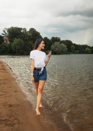 Foto de Joven alegre con el pelo largo camina por el lago descalzo. Mujer escuchando música.Chocando en la orilla. - Imagen libre de derechos