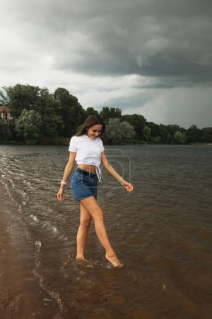 Téléchargez les photos : Joyeux jeune femme aux cheveux longs marche au bord du lac pieds nus. Éclaboussures sur le rivage. - en image libre de droit