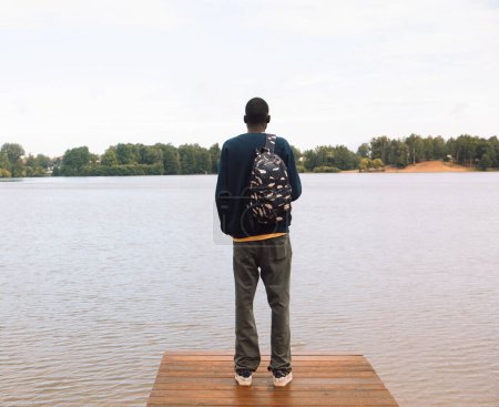 Foto de Un joven negro en ropa deportiva con una mochila camina cerca del lago. Vista trasera. - Imagen libre de derechos