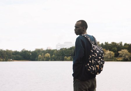 Foto de Un joven negro en ropa deportiva con una mochila camina cerca del lago. Concepto de estilo de vida. Día de verano. - Imagen libre de derechos