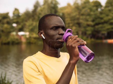 Foto de Joven hombre deportivo afroamericano guapo cansado y sediento después de hacer ejercicio sosteniendo botella de agua potable. Concepto de estilo de vida. - Imagen libre de derechos