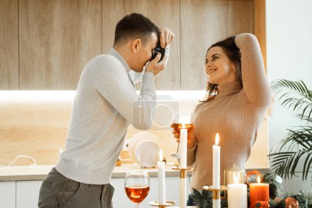 Photo for Young happy couple celebrating christmas, holding glasses of wine, man taking photo of his girlfriend. Lifestyle concept. - Royalty Free Image