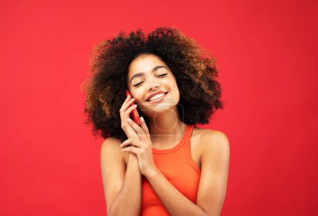 Foto de Retrato de una joven afro-americana emocionada hablando por teléfono móvil sobre fondo rojo. - Imagen libre de derechos