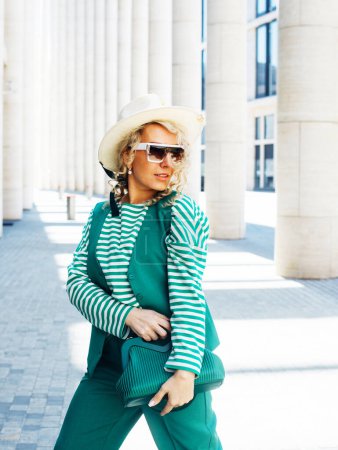 Photo for Smiling Woman Outdoor Portrait. Curly blonde hair fashion model wears stylish clothes, green leather handbag and sunglasses. Female fashion. Sunny day. Close up. City lifestyle. - Royalty Free Image