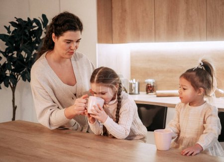 Foto de Dos niñas hermanas están tomando té con su madre en la cocina. Concepto de estilo de vida y personas. - Imagen libre de derechos