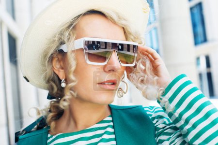 Foto de Retrato de una mujer adulta con el pelo rubio rizado con gafas de sol y sombrero. Retrato de la ciudad de cerca. - Imagen libre de derechos