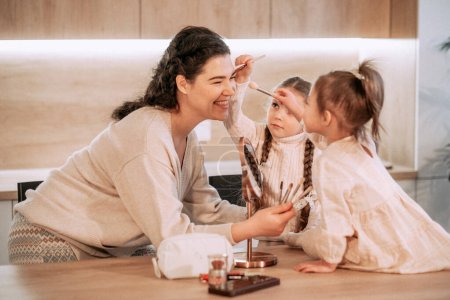 Foto de Dos hermanas niñas juegan con su madre usando cosméticos. Maquillaje juego. Feliz tiempo en casa. Concepto de estilo de vida. - Imagen libre de derechos