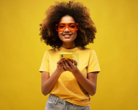 Photo for Portrait of lovely ethnic woman holds modern mobile phone, uses electronic device on surfing web, looks positively at camera, connected to wireless internet, wears yellow shirt and big sunglasses - Royalty Free Image